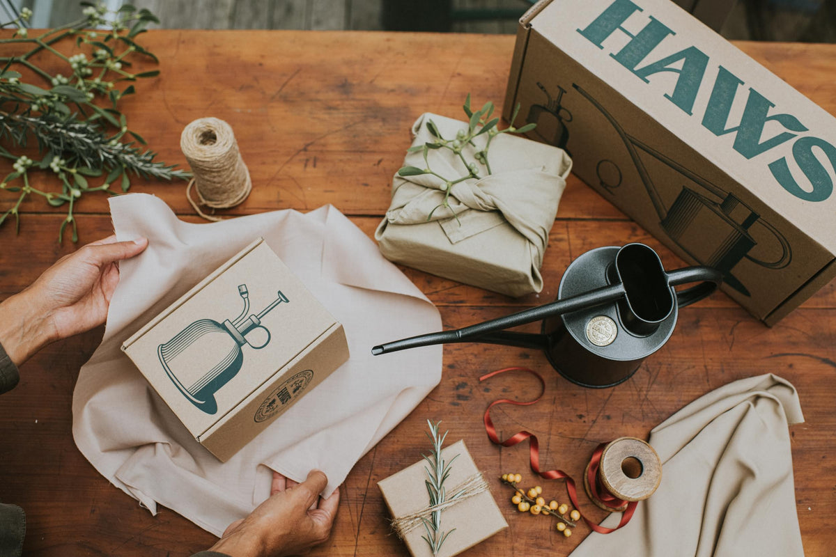 A styled flat-lay of a HAWS mug gift set on a rustic wooden table. The set includes a brown cardboard box with a teal HAWS logo, a matching mug box, a dark gray ceramic mug, a beige linen cloth, twine, dried herbs, and a small wrapped gift.