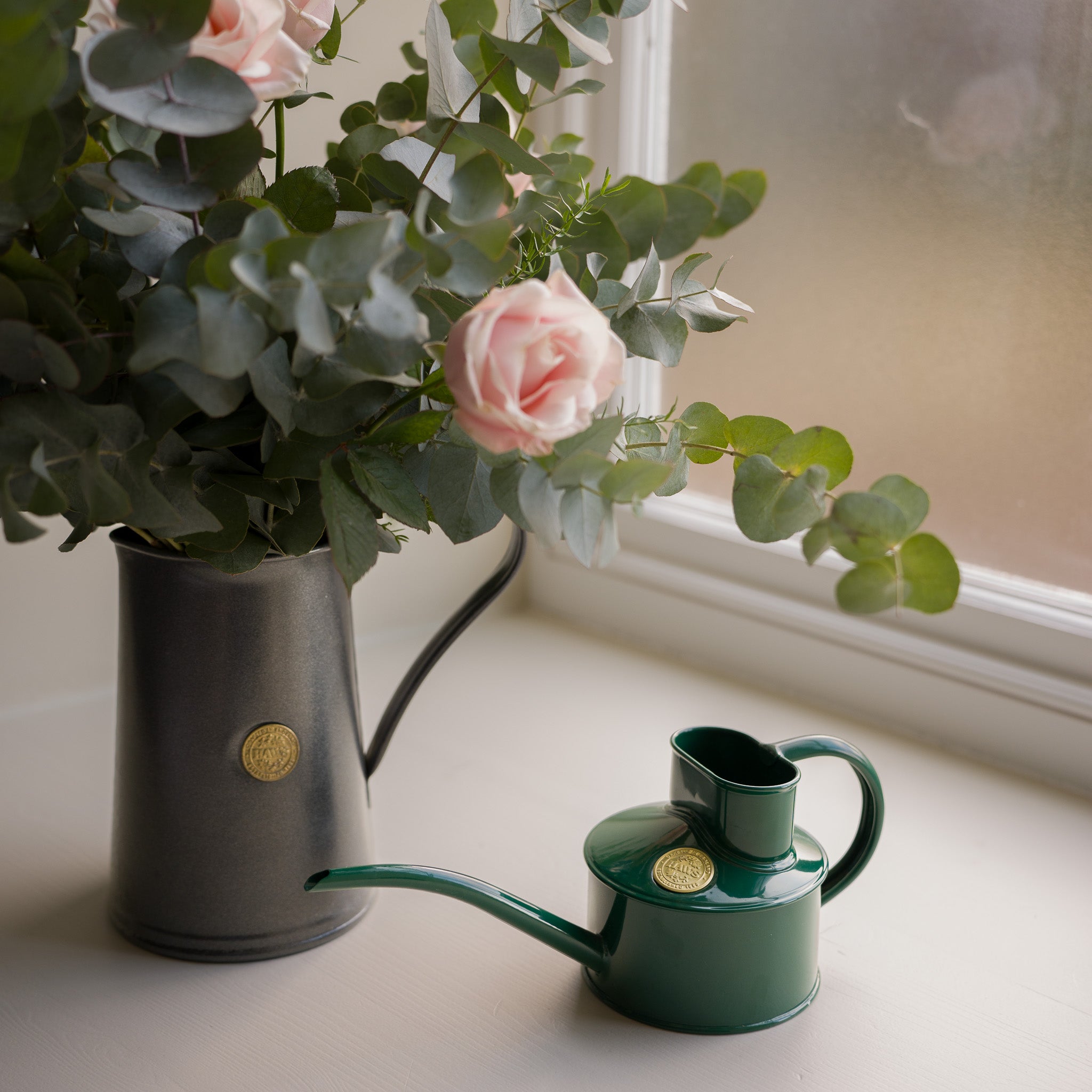 Small green Haws watering can next to a black Haws jug with pink roses and greenery on a windowsill.